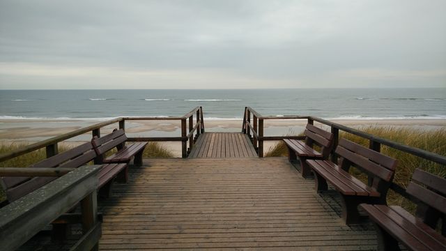 A wooden pathway with benches leads to a serene beach, overlooking a calm ocean under a cloudy sky. Great for travel blogs, tourism promotions, relaxation and meditation content, and nature-related articles.