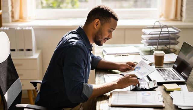 Man reviewing financial charts at sunlit home office desk using calculator and laptop