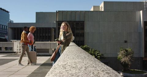 Sunlit urban travelers with luggage, woman leaning on concrete railing using smartphone