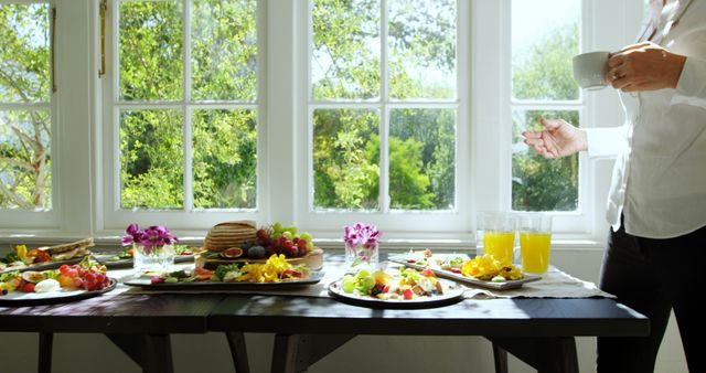 Woman stands by sunny window enjoying delicious breakfast spread. Various fresh and vibrant foods including fruits, juice, pancakes, and salads populate the table, indicating a healthy and delightful start to the day. Ideal for illustrating wellness, mornings, health food, dining experiences, and domestic environments.