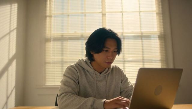 Young man focusing on laptop in sunlit room with blinds, remote work and study resource