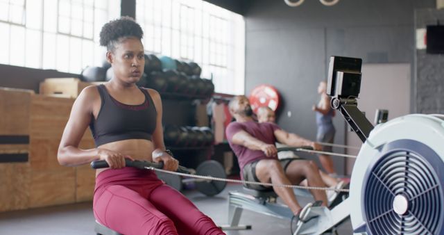 Young woman training intensely on a rowing machine in a gym. She is joined by other individuals also using rowing machines in the background. This image can be used for fitness-related topics, gym advertisements, online and offline training programs, or articles on health and wellness.