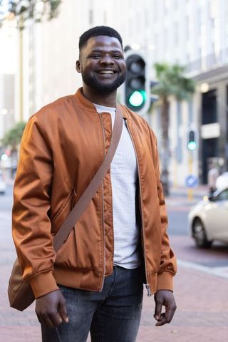 Smiling african american man standing on city sidewalk wearing brown bomber jacket