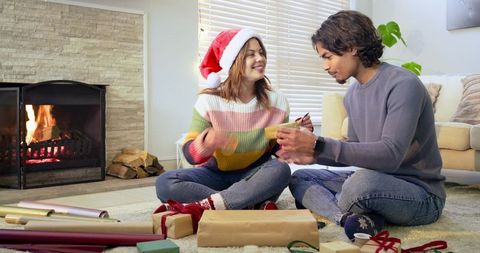 Couple wrapping christmas gifts by fireplace woman wearing santa hat cozy home