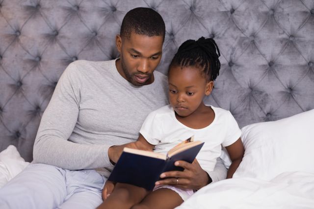 Father reading a book to his daughter while sitting on a bed at home. Ideal for use in parenting blogs, educational materials, family-oriented advertisements, and articles about father-daughter relationships and bedtime routines.