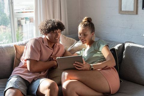 Sunlit young couple relaxing on sofa browsing tablet together, home lifestyle
