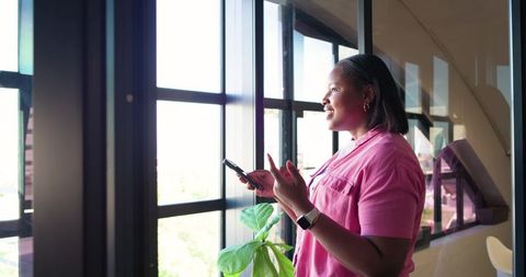African woman standing by office window holding smartphone, gesturing and smiling in sunlit workspac