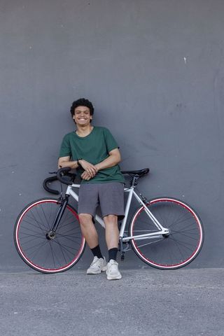 Young man leaning against wall with white road bike and red rims, smiling urban cyclist