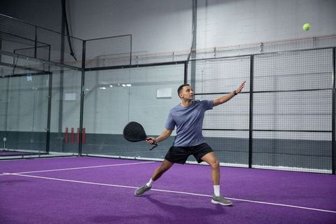 Male padel player lunging to hit ball on purple modern indoor court with glass panels and fence