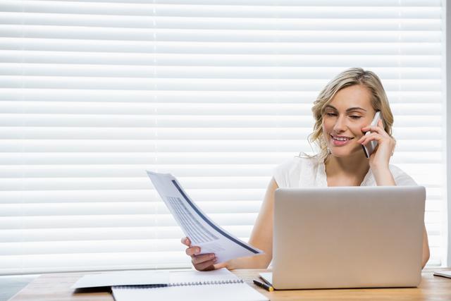 This image shows a woman multitasking by talking on a mobile phone while holding a document and working on a laptop at home. Ideal for illustrating concepts of remote work, business communication, productivity, and professional women in a casual home office setting.