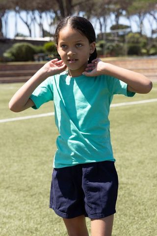 Schoolgirl adjusting necklace on sunlit turf field wearing turquoise tee and navy shorts