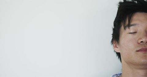 Resting asian man tilting head with closed eyes against pale wall minimal serene portrait