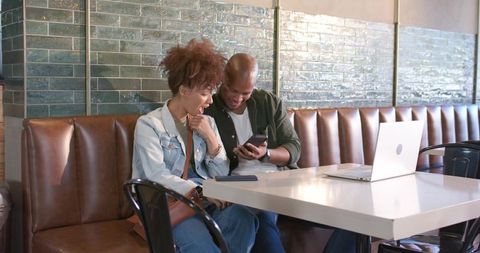 African american couple sharing smartphone and working on laptop in sunlit cafe booth