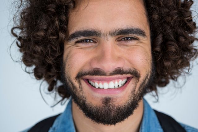 This image shows a close-up of a young male photographer with curly hair and a beard, smiling warmly. He is wearing a casual denim shirt and appears to be in a studio setting. This image can be used for promoting photography services, lifestyle blogs, or advertisements focusing on happiness and creativity.