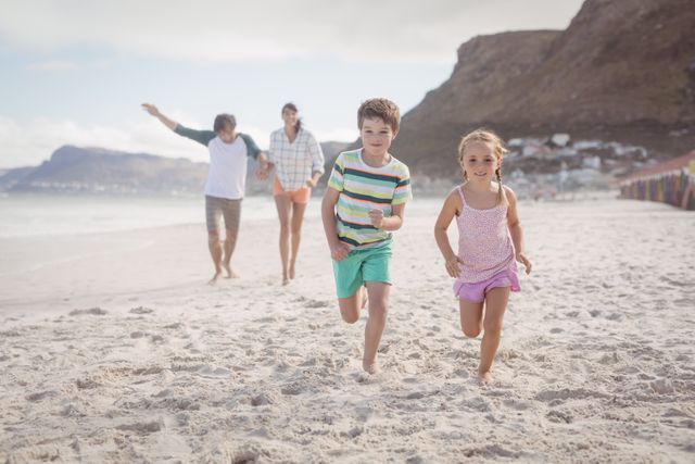 Siblings are running on the sandy beach with their parents in the background. The children are smiling and enjoying their time outdoors. This image is perfect for promoting family vacations, summer activities, and outdoor fun. It can be used in travel brochures, family-oriented advertisements, and websites focusing on family bonding and leisure activities.