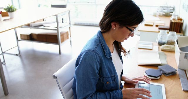 Young female professional focused on working on a tablet in a modern office. She appears to be engaged with her task, with various office supplies around her on the desk. Ideal for use in articles or marketing materials about modern workplaces, remote work, productivity, technology in business, or women in tech.