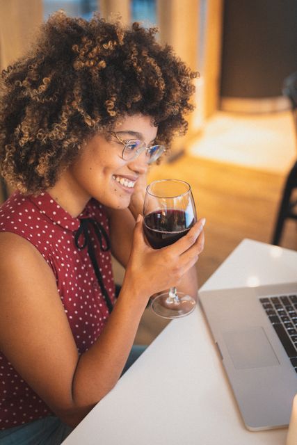 Biracial woman with curly hair and glasses smiling while having a video call on her laptop, holding a glass of red wine. Ideal for use in articles or advertisements about online communication, socializing, technology in everyday life, or leisure activities. Perfect for illustrating modern social interactions and the use of technology in maintaining relationships.