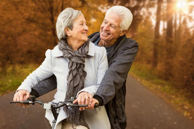 Senior couple enjoying a bicycle ride in an autumn park, surrounded by colorful trees. They are smiling and looking at each other, showcasing happiness and love. Ideal for use in advertisements, health and wellness campaigns, retirement planning materials, and articles about active aging and outdoor activities.