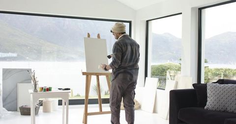 Indian artist painting at easel in bright lakeview studio with mountain backdrop
