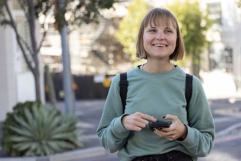 Smiling young woman holding smartphone on sunny urban sidewalk with backpack