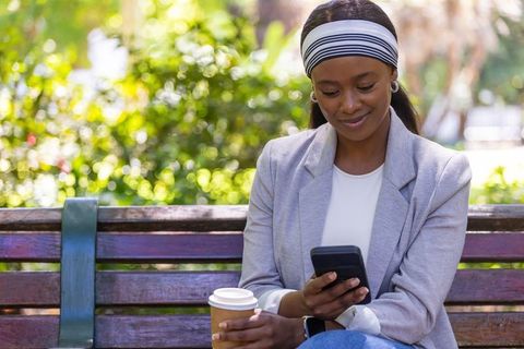 African woman sitting on park bench checking phone while holding coffee cup