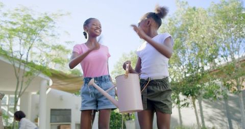 Smiling girls holding watering can enjoying sunny backyard gardening and playful bonding