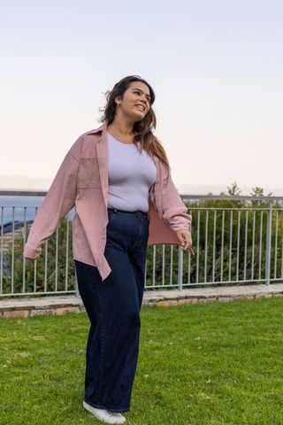 Young woman walking and smiling on waterfront lawn wearing pink overshirt and denim jeans