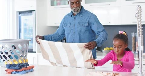 African american grandfather teaching granddaughter folding laundry at modern kitchen island