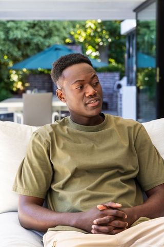 African american man sitting on sunroom couch wearing olive green t-shirt looking right