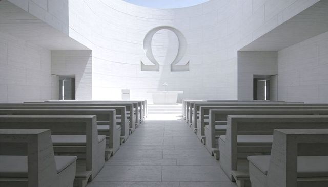 Showcasing minimalist white chapel with carved omega above central altar, symmetrical stone pews