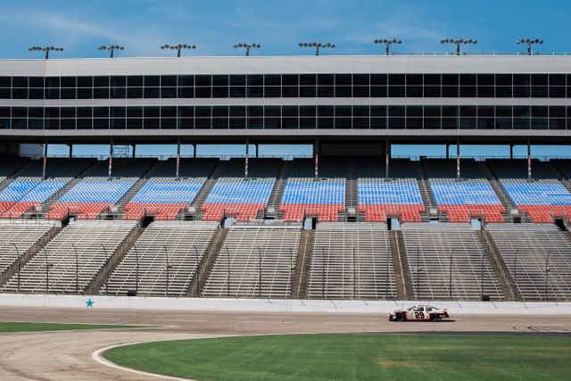 A race car driving on a racetrack with empty stadium seating. Ideal for themes involving motorsports, racing events, automotive industry, speed, and empty venues. Useful for advertisements, blog posts, and promotional materials about races or sports events.