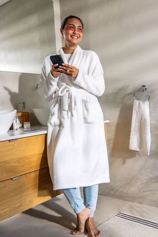 Smiling asian woman in white robe using smartphone during relaxed morning skincare routine