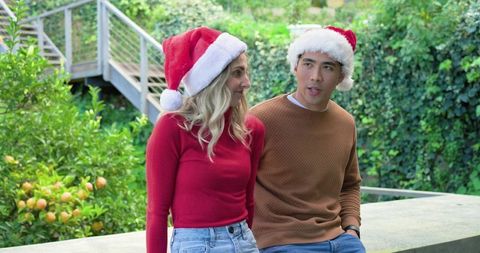 Festive outdoor couple wearing santa hats sitting on backyard ledge beside citrus tree