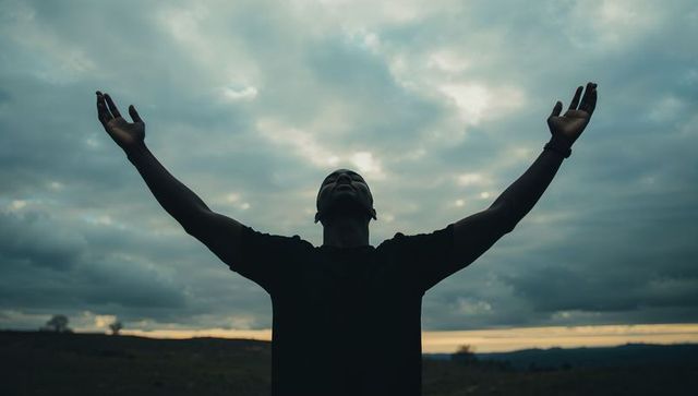 Male silhouette raising arms at moody sunset over rural plain conveying solitude