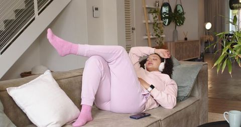 Relaxed african american woman on sofa in pink loungewear listening to music with headphones