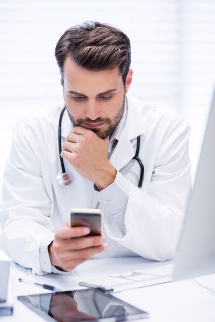 Male doctor in white coat with stethoscope using mobile phone in clinic. Ideal for illustrating modern healthcare, medical technology, communication in healthcare, and professional medical environments.
