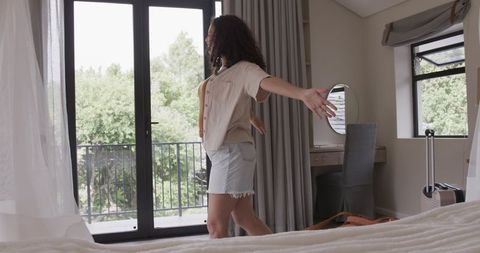 African american woman barefoot stretching in bright hotel bedroom by balcony with luggage