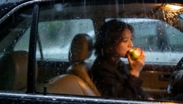 Woman biting apple inside rain-soaked car window, reflective mood, urban commute