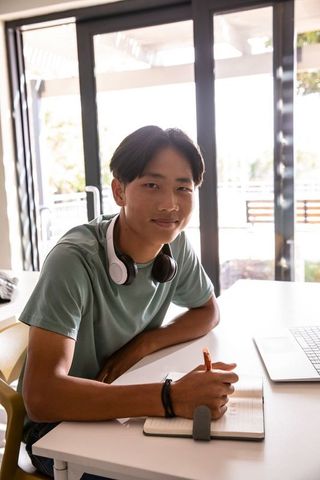 Asian teenager studying at desk with laptop and headphones in bright modern home workspace