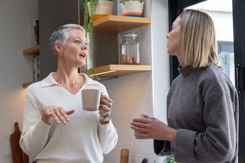 Senior woman and friend chatting over coffee in sunlit cozy kitchen holding ceramic mugs