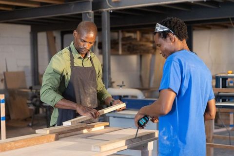 African american mentor guiding apprentice sanding and drilling wood in carpentry workshop