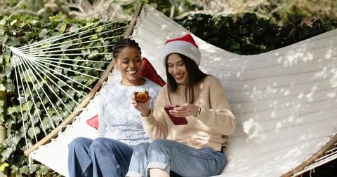 Diverse women sharing phone and drink on hammock while wearing santa hat in sunny backyard
