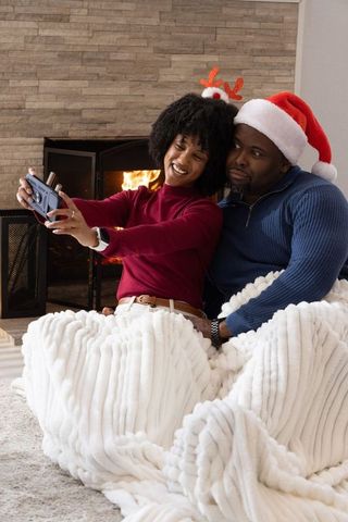 African american couple taking festive selfie by fireplace wrapped in cozy blanket