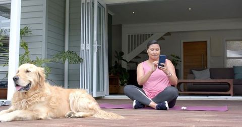 Woman sitting cross-legged on deck with golden retriever, scrolling smartphone and relaxing