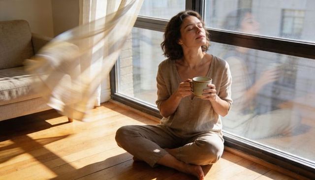 Morning calm woman sitting in loungewear holding mug by window with billowing curtain