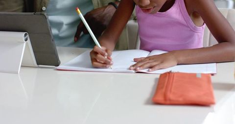 African american mentor guiding young girl writing homework using tablet and notebook