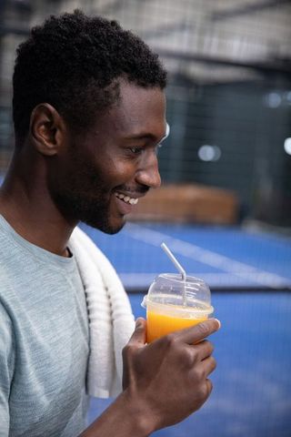 Smiling african american man hydrating with orange juice after tennis court workout