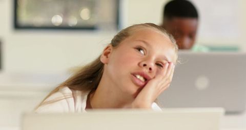 Daydreaming preteen girl in classroom with classmate working on laptop, learning distraction