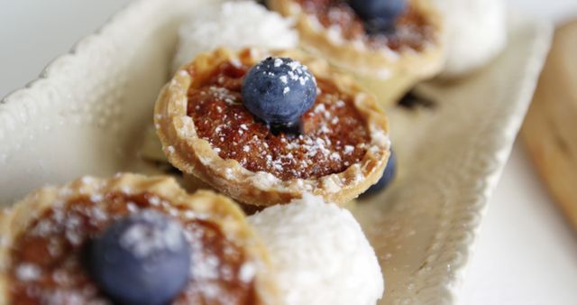 This image features a close-up view of gourmet mini fruit tarts adorned with fresh blueberries and a dusting of powdered sugar, accompanied by dollops of cream. Perfect for use in culinary blogs, dessert menus, bakery advertisements, and food and lifestyle magazines to showcase elegant sweets and baking artistry.