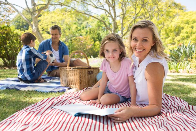 This image depicts a happy family enjoying a sunny day in the park. The mother and daughter are sitting on a striped blanket, reading a book, while the father and son are engaging with a football in the background. The lush greenery and bright atmosphere suggest a fun, relaxing family outing. This photo is ideal for use in advertisements, blog posts, and articles related to family, outdoor activities, leisure time, and bonding.